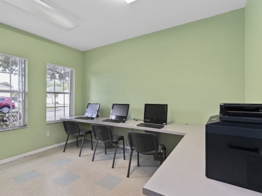 a computer room with three computers on a long desk with chairs at The Groves at Wimauma Apartments , Wimauma, Florida
