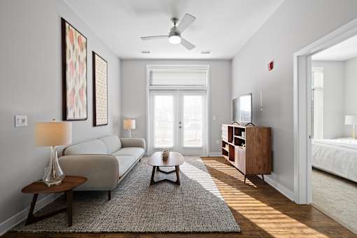 A living room with double doors to a patio providing natural light, hardwood-style flooring, a ceiling fan, and access to a bedroom.