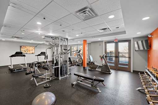 A spacious fitness center at New Kent Apartments in West Chester, PA, featuring cardio and weight equipment, recessed lighting, large windows, a TV, and double doors opening to the outside