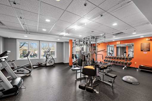 A fully-equipped fitness center at New Kent Apartments in West Chester, PA, featuring weight and cardio machines, large mirrors, and ample natural light from the windows