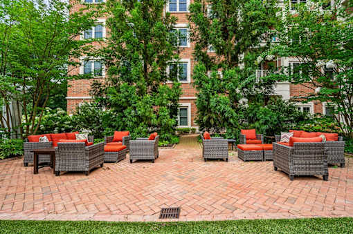 A patio with red cushions and grey wicker furniture is surrounded by trees and a brick building.