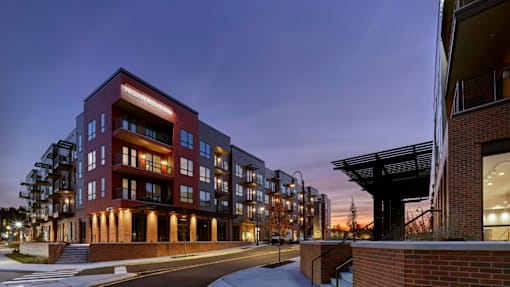 A corner street view of River Station's apartment buildings at sunset with trees and the capitol building in the background.