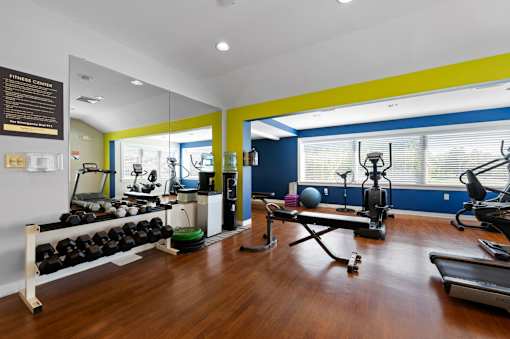 A state-of-the-art fitness center with free weights, weight lifting benches, cardio machines, hardwood-style flooring, recessed lighting, and large windows.