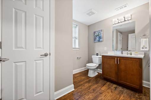 A white door is on the left of a bathroom with a toilet, sink and mirror.