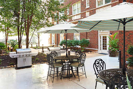 Courtyard with concrete pad, black iron table with chairs, white umbrella, gas grill and trees in the background at Residences at Forest Park, Saint Louis, MO