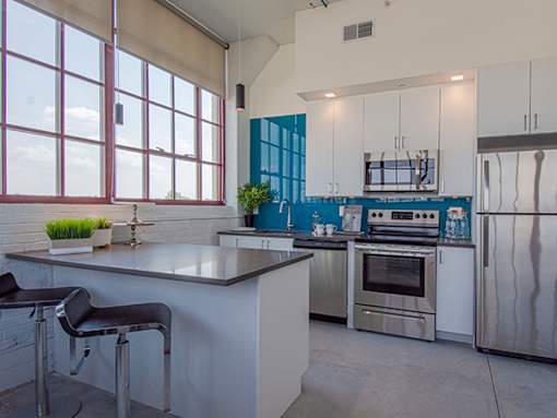 Steelcote Lofts kitchen with Steelcote blue backsplash, gray quartz countertops, stainless steel appliances, white cabinetry, kitchen island and polished concrete floors at Steelcote Square, St. Louis