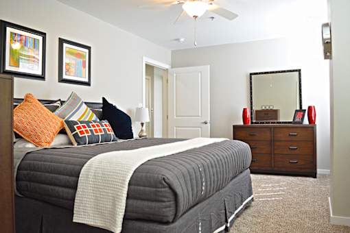 bedroom with light carpeting, bed with gray duvet, brown dresser with mirror at Residences at Forest Park, Saint Louis, MO, 63108