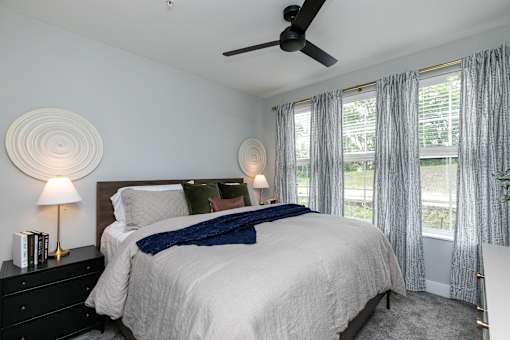 A bedroom with a large bed, a ceiling fan, and a window with curtains at The Cunningham Apartments, West Des Moines, Iowa