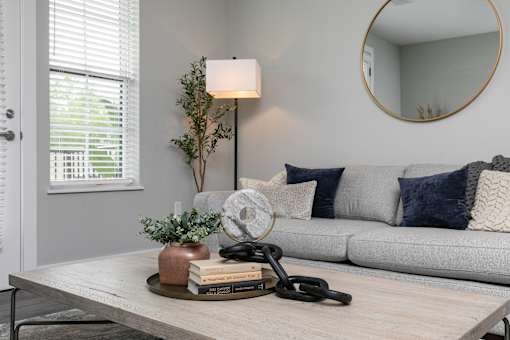 A living room with a grey couch, a wooden coffee table, and a round mirror on the wall. at The Cunningham Apartments, Apartments, West Des Moines, IA