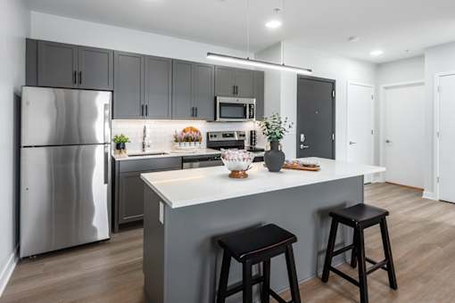 model kitchen with stainless steel appliances, plank flooring, kitchen island, white quartz countertops, gray cabinetry and white tile backsplash at Steelcote Square, St. Louis, 63103