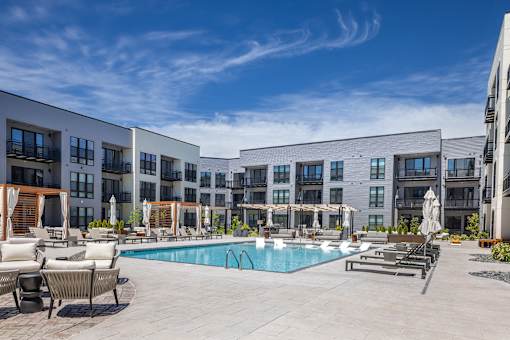 A large swimming pool surrounded by lounge chairs and umbrellas in front of a modern apartment complex.
