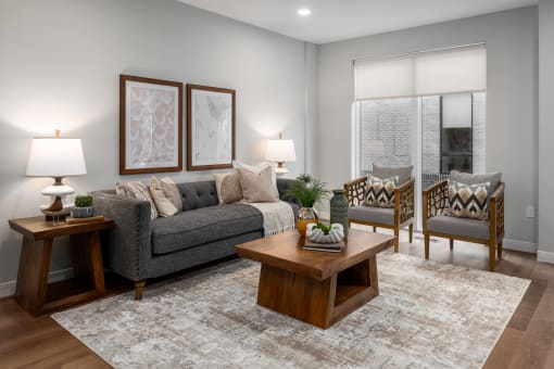 Living room with rich plank flooring, oversized windows with custom shades, full size sofa, two arm chairs, coffee table and end table at The Flats & Terraces at Wildhorse Village, Missouri, 63005