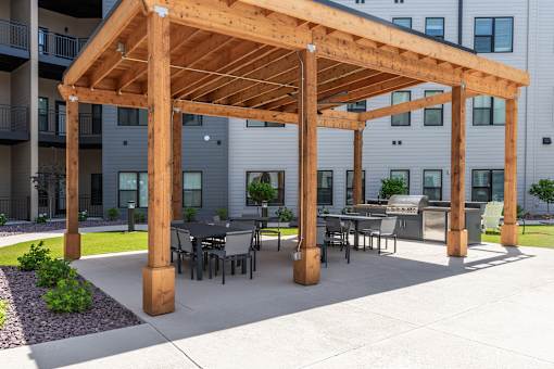 A wooden pergola with a table and chairs is in front of a building.