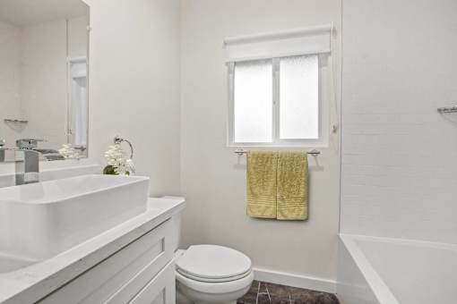 A white bathroom with a toilet, sink, and a window with a yellow towel hanging on it.
