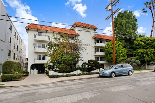 A blue car is parked on the street in front of a white building with a red tile roof.