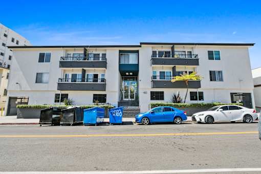 A blue car is parked in front of a white building with balconies.
