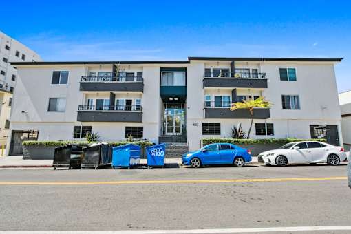 A blue car is parked in front of a white building with balconies.