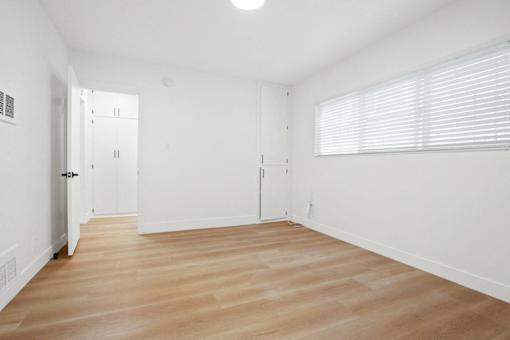 A room with white walls and wooden flooring with a door on the left and a window with blinds on the right.