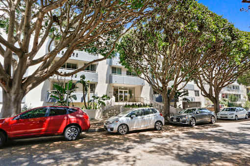 A red car is parked in front of a white building with trees.