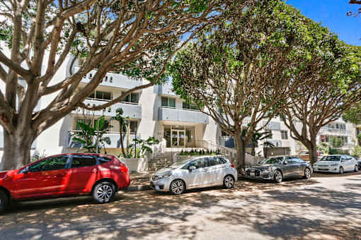 A red car is parked in front of a white building with trees.