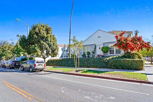 A street view with cars parked on the side of the road and a white house with a red flower bush in front.