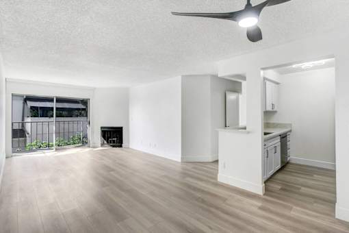 A spacious room with a ceiling fan and light, hardwood floors, and a kitchenette with white cabinets.