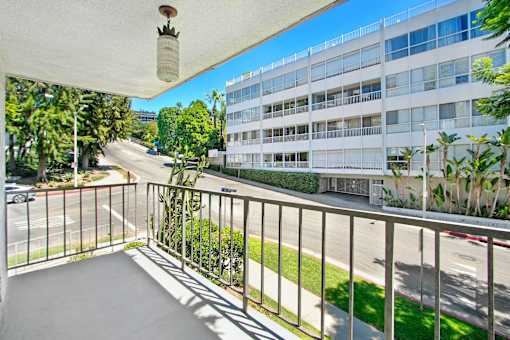 A balcony with a white railing and a hanging light fixture overlooks a street with a white building in the background.