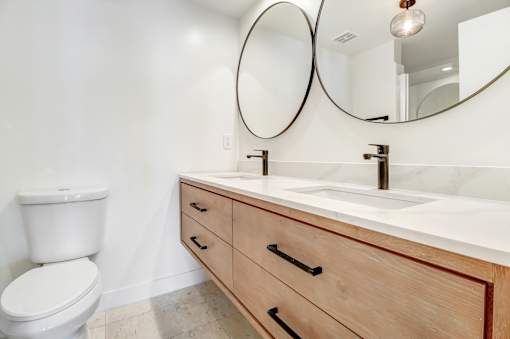 A white toilet sits next to a sink with a marble countertop and two round mirrors above it.