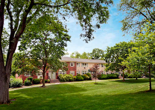 A large red brick building with a white door and windows is surrounded by green trees and grass. at Rivercrest Apartments, New Jersey, 08854