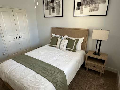 A bed with a green blanket and pillows is in the foreground with a lamp and two pictures on the wall.