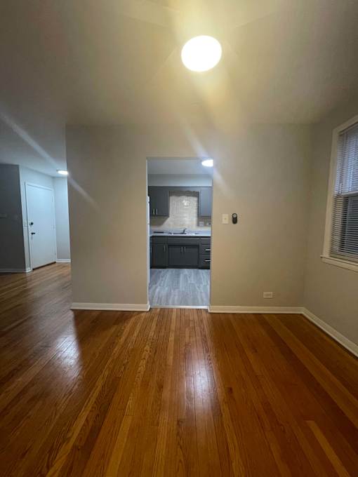 A room with wooden floors and a kitchen visible through the door.
