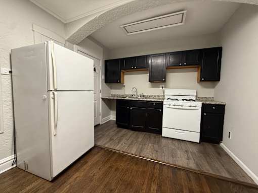 A white refrigerator stands in a kitchen with black cabinets and a white stove.