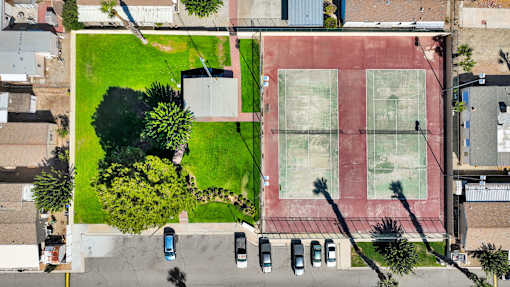 A tennis court is surrounded by a grassy area and a parking lot.