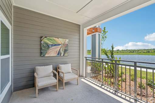 Two chairs are on a porch with a painting and a view of a lake.