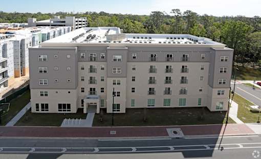 A large, modern building with a flat roof and multiple windows is surrounded by a parking lot and other buildings at Arbours at Tumblin Creek ~ Active 55+ Adult Community, Gainesville, FL