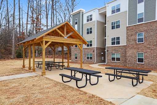A wooden pavilion with picnic tables is situated in front of a brick building.