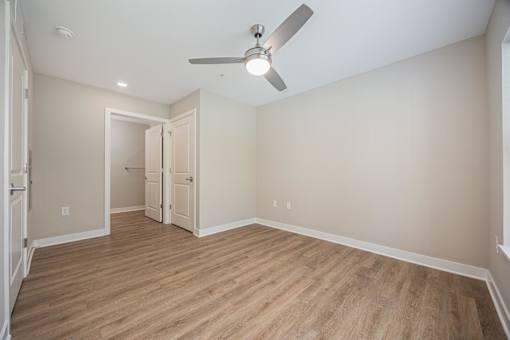 A room with a ceiling fan and light fixture, hardwood floors, and a doorway leading to another room.