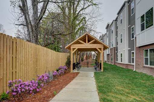 A wooden gazebo is situated in a grassy area between two buildings.