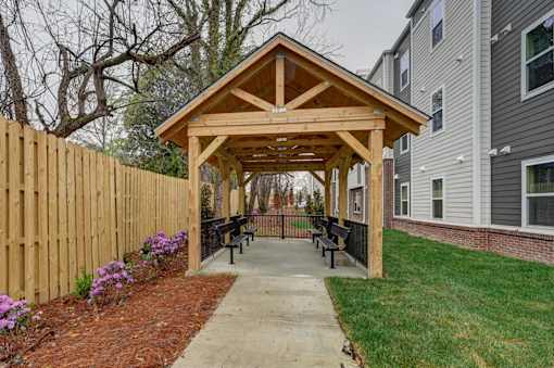 A wooden gazebo with a roof is surrounded by a wooden fence and a concrete walkway.