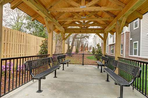 A wooden pergola with black benches is in front of a fence and houses.