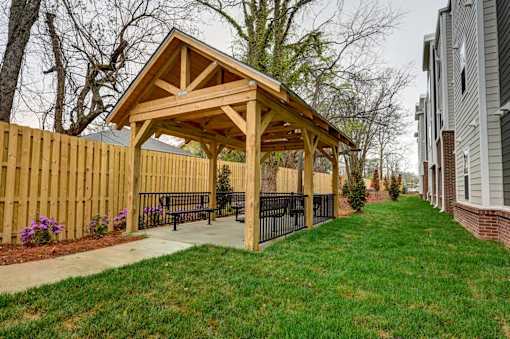 A wooden pavilion with a black fence and benches is surrounded by a grassy area.