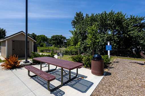A picnic table with a red top and two benches is in the foreground of a sunny outdoor area.