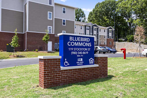 A sign for Bluebird Commons apartment complex stands in front of a brick building.