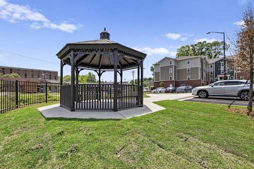 A gazebo is situated in a grassy area with a black fence and a car parked in the background.
