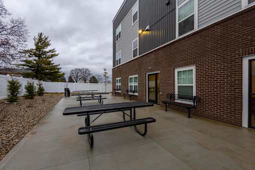 A series of black picnic tables are lined up on a concrete patio outside of a brick building.