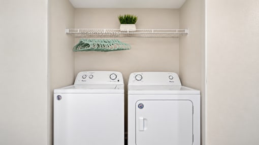 Apartment laundry closet with a side-by-side washer and dryer and overhead wire shelving at Canterbury Apartments in Myrtle Beach, SC.