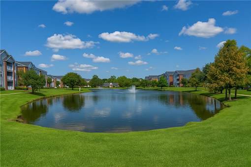 A serene landscape featuring a pond, a fountain, and a row of buildings.