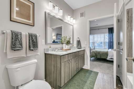 A bathroom with a white toilet, a mirror, and a bedroom in the background.