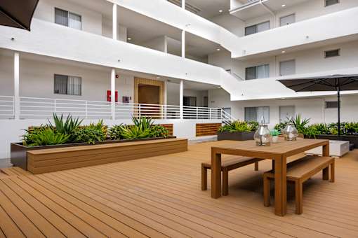 A wooden table with benches is in the foreground of a modern building with white balconies.