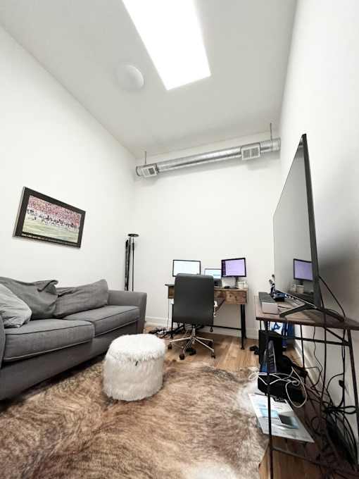 A room with a grey couch, a white ottoman, a desk with a computer and a monitor, and a picture on the wall.